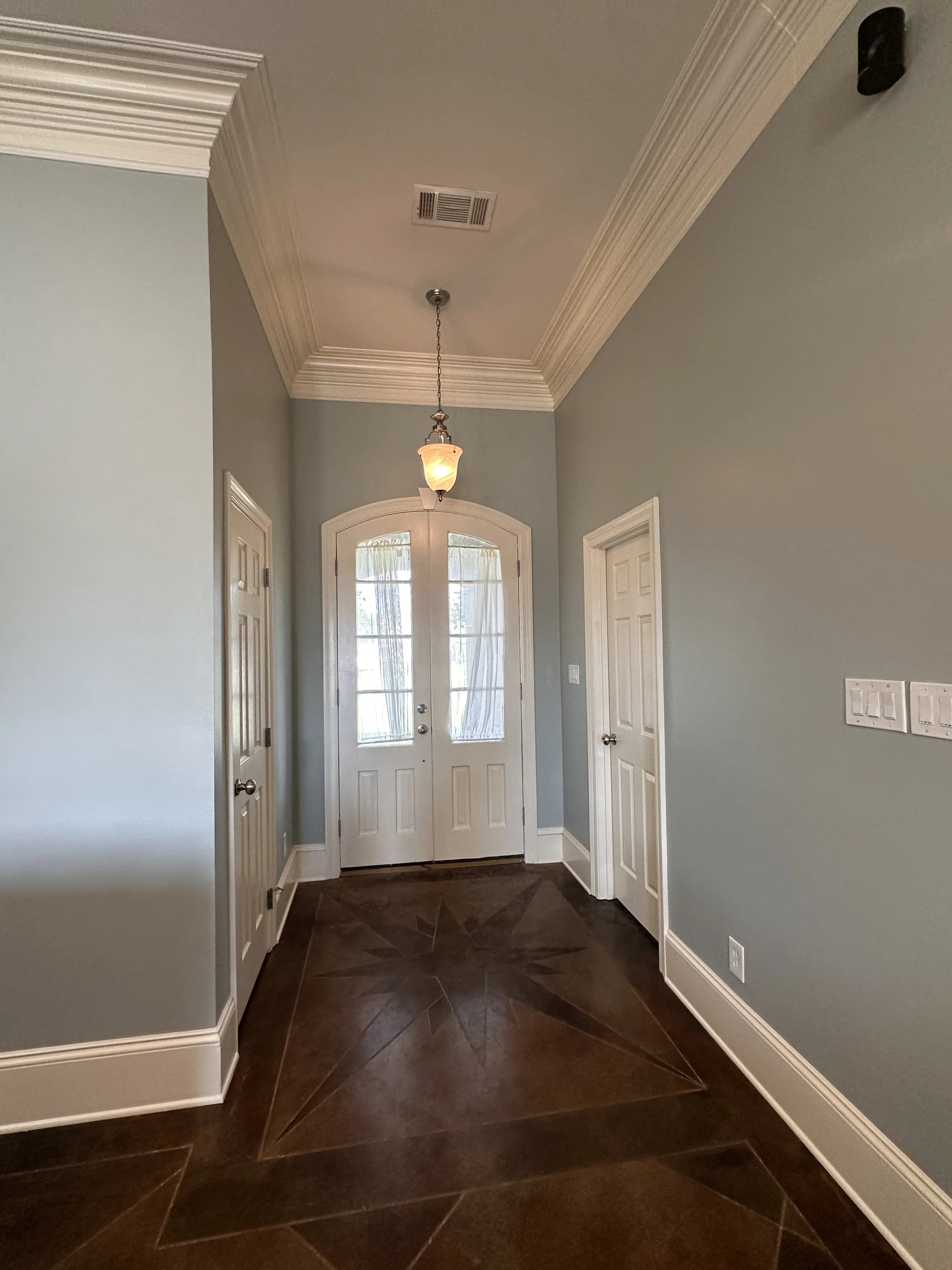 A well-lit hallway with gray walls, white trim, and a dark tiled floor featuring a star pattern, leading to arched French doors.