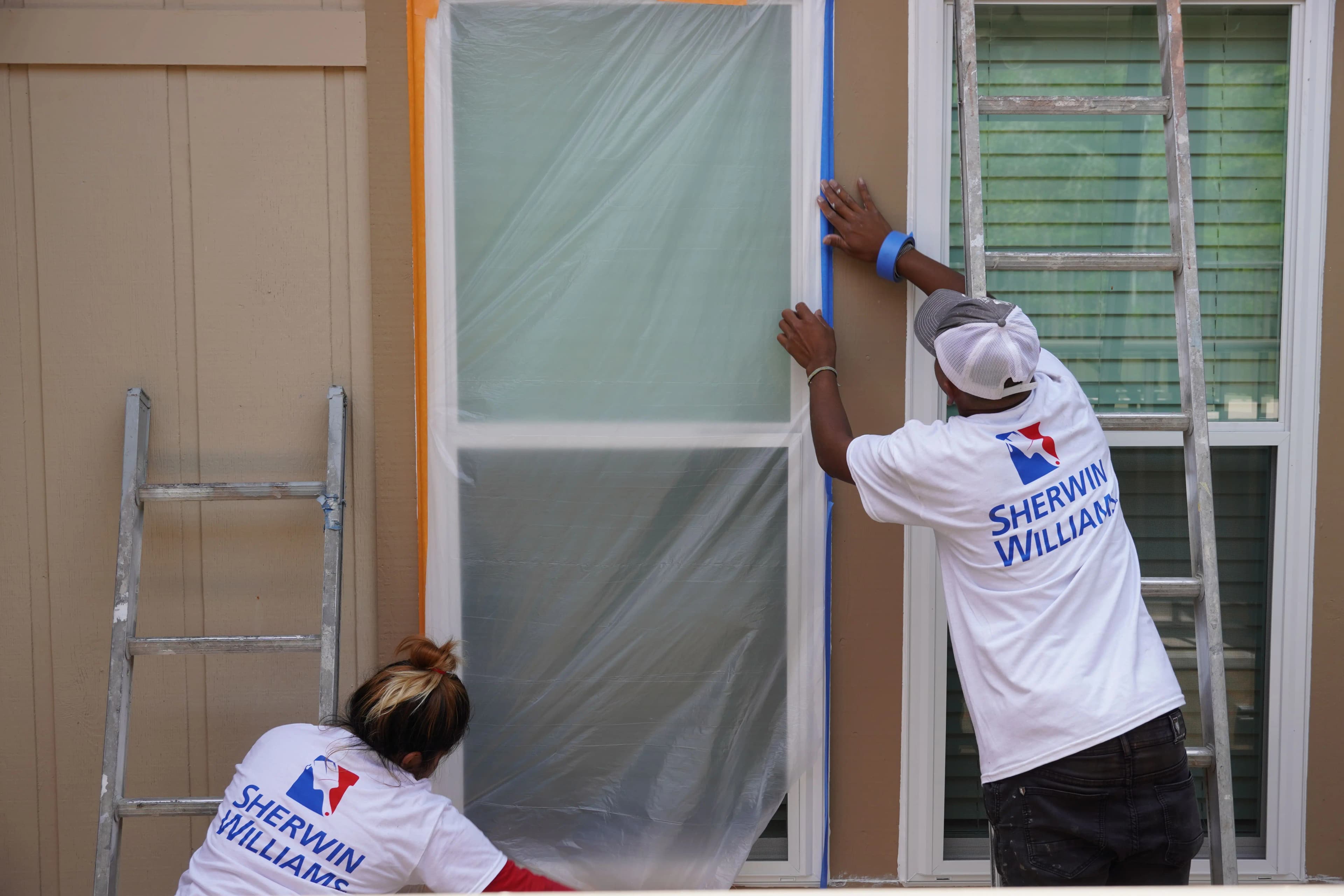Two Sherwin-Williams painters carefully tape off a window with plastic sheeting before painting a house exterior.