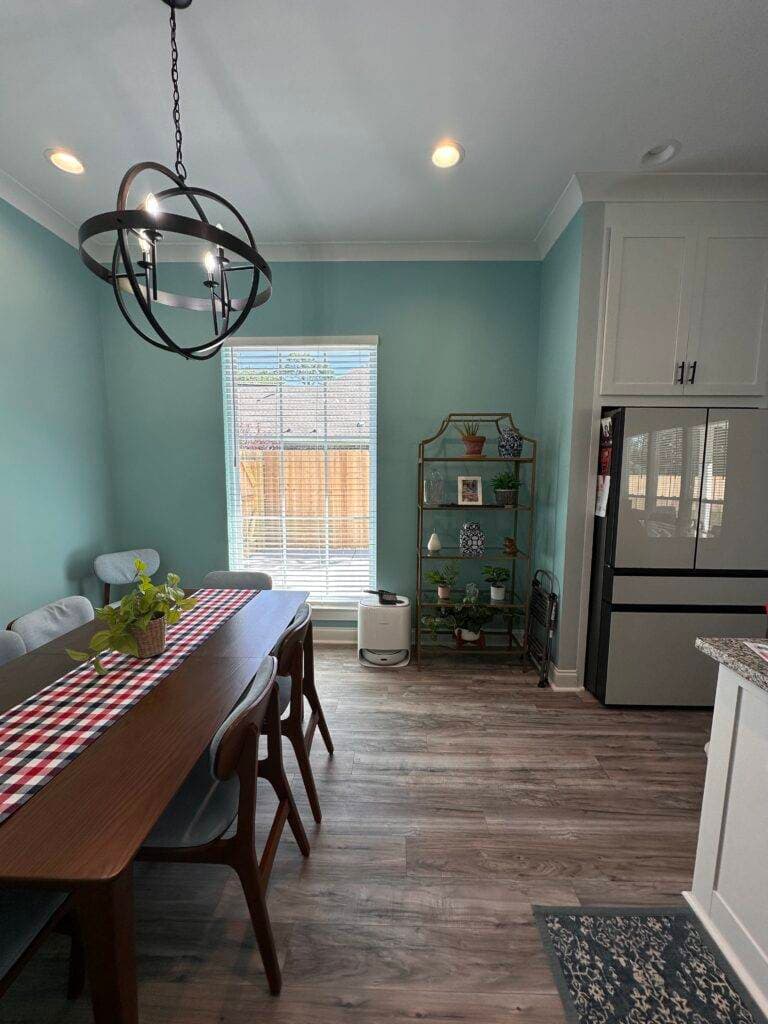 A contemporary dining area with a wooden table, modern pendant light, and light blue walls adjacent to a kitchen.
