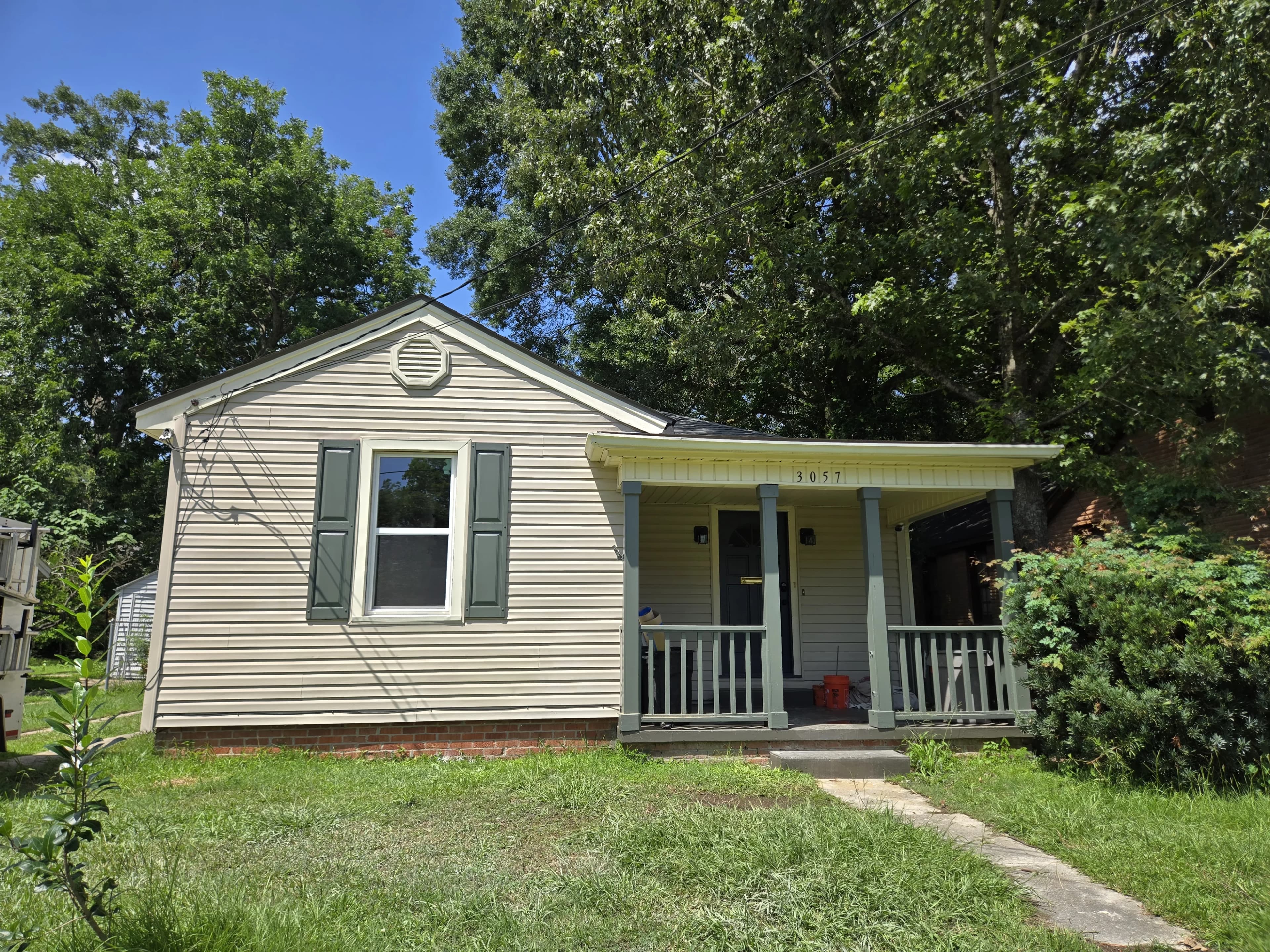 A beige single-story house with a front porch and green shutters, surrounded by lush greenery under a clear blue sky.