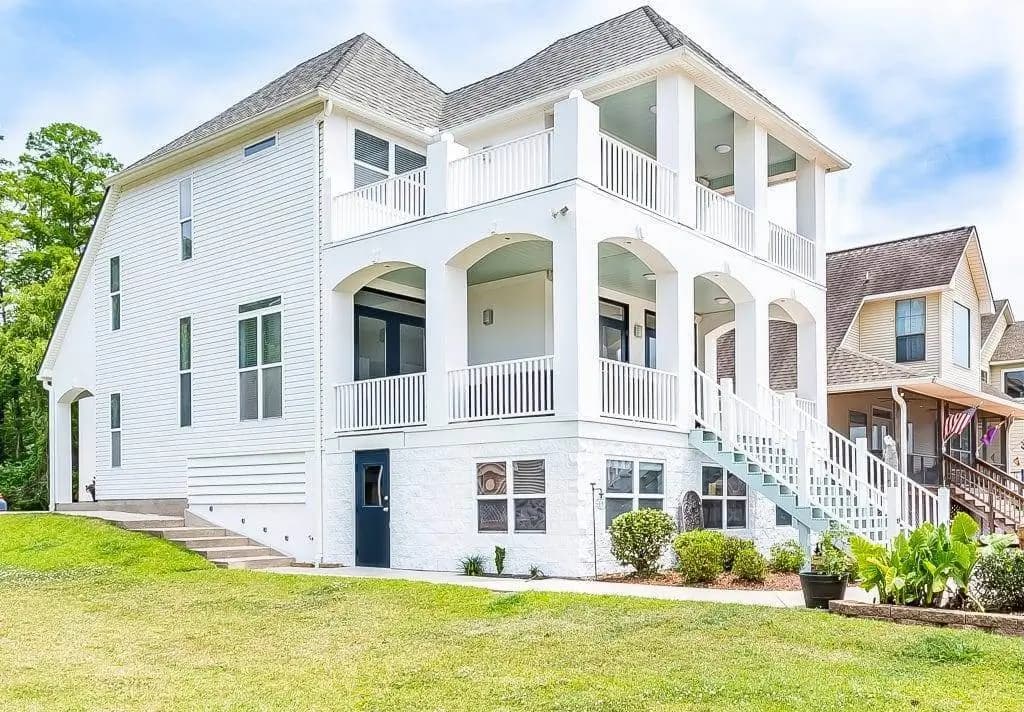 A white two-story house with balconies and a blue front door, set on a grassy lawn under a partly cloudy sky.